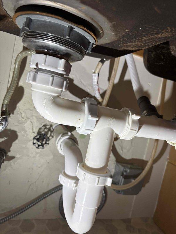 Responded to warranty callback for kitchen stack leak affecting multiple floors. Upon inspection, found water accumulation under sink cabinet following previous service visit. Inspected PVC drain assembly, P-trap configuration, garbage disposal connections, and all slip-joint fittings for proper sealing and alignment. Documented existing plumbing configuration including white PVC trap assembly, supply lines, shut-off valves, and disposal unit connections to verify installation integrity and identify source of continued leakage.