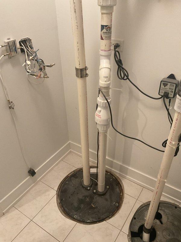 Responded to service call for residential ejector pump reported as not cycling. Performed diagnostic inspection of the sump pump system located in basement utility area. Assessed pump basin, discharge piping configuration including white PVC vertical and horizontal runs, electrical connections, and standing water level in basin. Documented existing plumbing installation with photos showing pump basin with cover displaced, dual vertical PVC discharge pipes, wall-mounted water heater with associated plumbing connections, and electrical supply components including outlet and transformer. Explained that replacement cost estimates would be provided on-site following complete system evaluation.