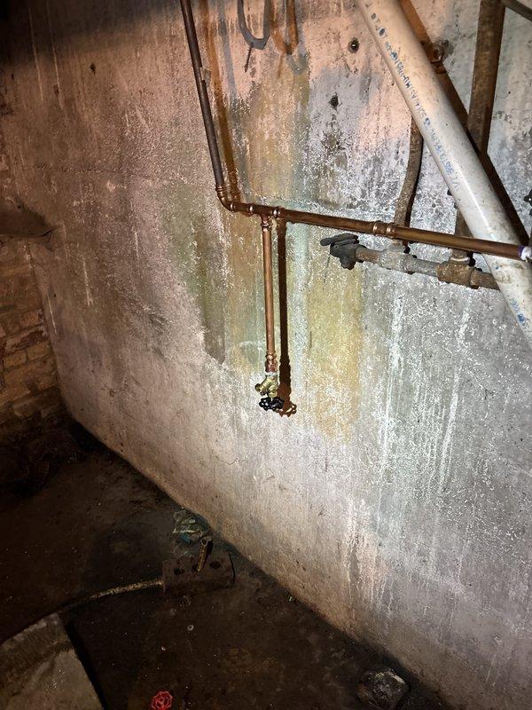 # Boiler Drain Installation and Plumbing System Repair

Completed warranty boiler drain installation in basement mechanical space with additional courtesy spot repair of aging copper piping system. Installed new brass boiler drain valve on existing vertical copper supply line, ensuring proper positioning and accessibility for future maintenance operations. 

Addressed deteriorating plumbing infrastructure showing significant corrosion and aging throughout the basement area. Repaired T-junction copper pipe configuration mounted on foundation wall, replacing corroded fittings and securing proper mounting brackets to ceiling joists. Evaluated extensive moisture damage and mineral deposits on basement walls indicating long-term water infiltration issues, which were noted for future monitoring.

Inspected horizontal copper supply lines running parallel to concrete ceiling alongside insulated gray piping. Verified all mounting straps and brackets were secure and properly supporting pipe runs