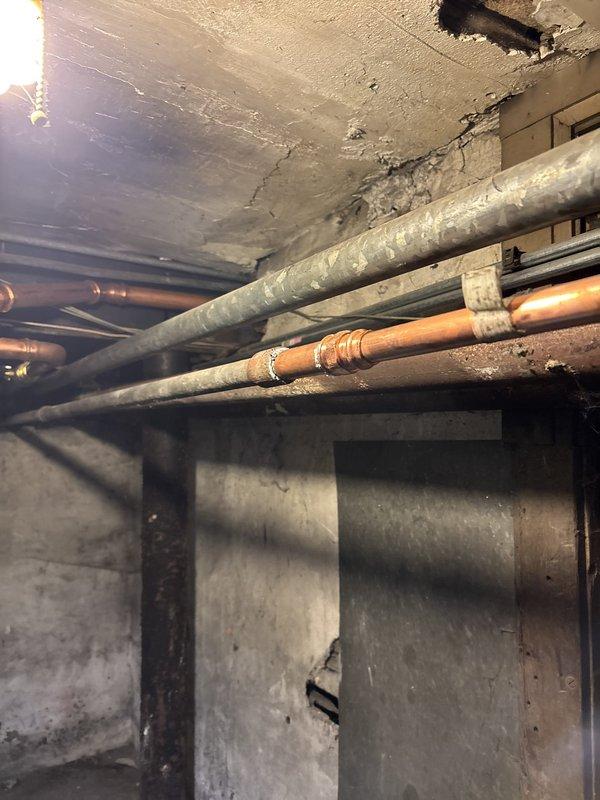 # Boiler Drain Installation and Plumbing System Repair

Completed warranty boiler drain installation in basement mechanical space with additional courtesy spot repair of aging copper piping system. Installed new brass boiler drain valve on existing vertical copper supply line, ensuring proper positioning and accessibility for future maintenance operations. 

Addressed deteriorating plumbing infrastructure showing significant corrosion and aging throughout the basement area. Repaired T-junction copper pipe configuration mounted on foundation wall, replacing corroded fittings and securing proper mounting brackets to ceiling joists. Evaluated extensive moisture damage and mineral deposits on basement walls indicating long-term water infiltration issues, which were noted for future monitoring.

Inspected horizontal copper supply lines running parallel to concrete ceiling alongside insulated gray piping. Verified all mounting straps and brackets were secure and properly supporting pipe runs