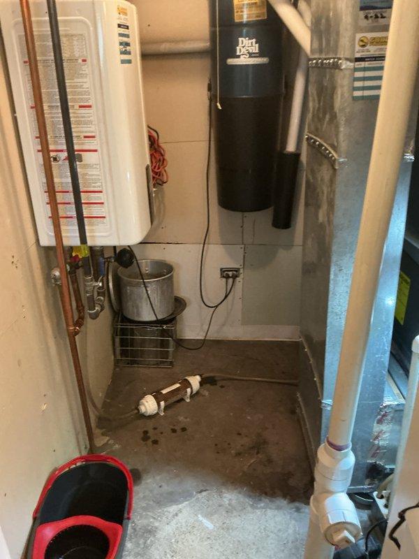 Responded to service call for tankless water heater that developed a leak following scheduled maintenance visit. Upon arrival, inspected white wall-mounted tankless unit and associated copper supply piping for source of water intrusion. Identified active leak condition requiring immediate attention. Placed collection bucket beneath unit to capture water discharge and prevent further damage to basement utility area. Documented equipment condition and leak location with photographs showing tankless heater installation, surrounding plumbing connections, PVC drainage system, and floor moisture patterns. Unit is a 2022 model requiring further diagnostic evaluation and repair to resolve leak issue and restore proper operation.