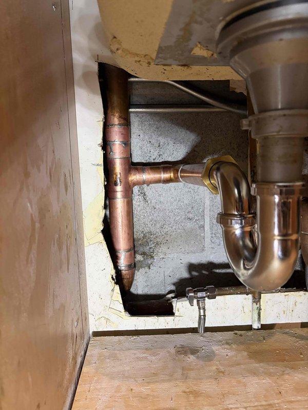 Performed plumbing repair at the kitchen sink, accessing the under-sink cavity and the wall cutout to service the copper supply lines, soldered joints, and fittings. Inspected and repaired the copper piping and connections, then verified the shutoff valve, braided flexible supply line, and chrome P-trap drain assembly were properly seated and secure, with the garbage disposal connection left intact.