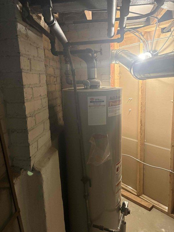 Evaluated the existing 20-year-old gas-fired tank water heater (A.O. Smith ProMax) and its gas supply, venting, and surrounding piping, then reviewed tank versus tankless options on site to determine the best replacement approach for the application. Inspected the exterior wall penetration and conduit/piping at the building's base to verify proper routing and sealing in preparation for the water heater installation.