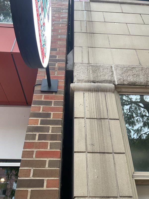 Performed a water leak locate to investigate reported moisture infiltrating the main entryway, suspected to originate from the unit porch drain. Visual inspection of the building exterior at the brick-to-limestone junction revealed significant water runoff staining and streaking below the projecting stone ledge, along with debris in the expansion joint, indicating exterior envelope water intrusion rather than a failure of the porch drain. Findings documented with photographs for repair planning and recommended sealing of the joint and ledge to prevent further water migration.