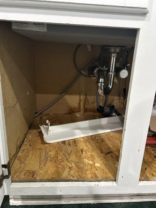 Inspected the dishwasher area for a reported leak and opened the adjacent wall cavity, exposing the copper supply lines and degraded insulation. Examined the sink base cabinet and drain assembly, finding long-term moisture damage with swollen, stained OSB flooring and water-streaked particleboard panels around the P-trap, tailpiece, and flexible supply line connections.