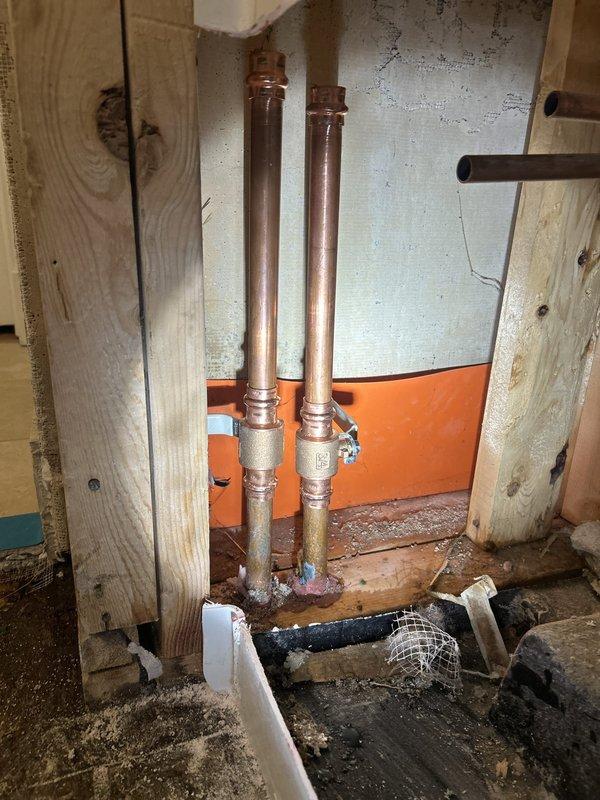 Performed plumbing piping installation work in a bathroom with the tub access panel opened, exposing the PVC drain assembly, and in an adjacent open wall cavity containing paired vertical copper supply lines with valve fittings. Inspected the copper connections and noted corrosion and flux residue at the lower fittings, then addressed the piping and fittings to restore proper connections with the subfloor and framing left accessible for follow-up work.
