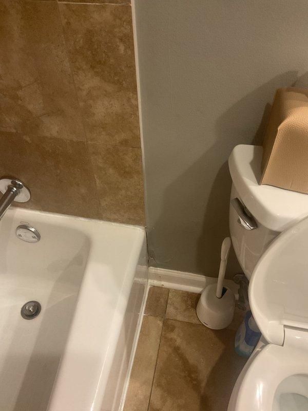 Performed a water leak locate investigation in response to reported moisture behind the bathroom wall, visually inspecting the tub surround, fixtures, and adjacent wall and floor areas. Documented evidence of moisture intrusion, including paint seam cracking at the wall-to-tub transition, staining on the floor tile near the tub and toilet, and ceiling discoloration in the adjacent room below, indicating a probable active leak in the tub-area supply or drain lines.