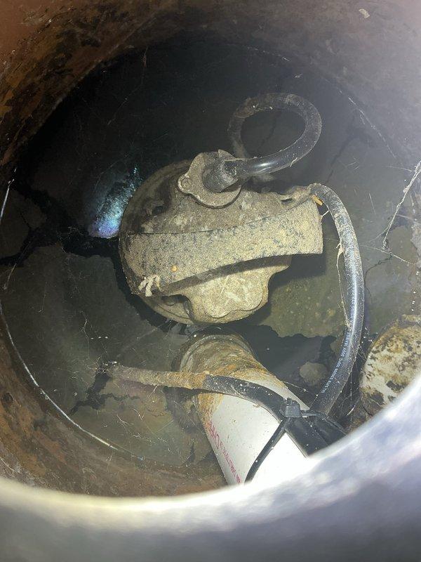 Inspected the ejector pump system in response to reports of excessive cycling during rain events. Located the sump basin beneath the exterior access lid and found a submersible pump heavily coated in sediment and grime, with a corroded basin, mineral and rust buildup on the discharge piping, and cracking and staining in the pit walls. The condition indicates likely infiltration and debris accumulation contributing to the frequent run times, and the basin and pump are recommended for cleaning and further evaluation.