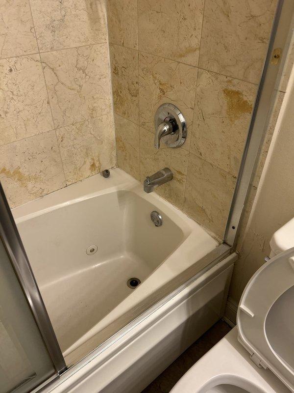 Performed a water leak locate investigation in response to a reported ceiling leak originating from an adjacent bathroom. Inspected the tub/shower alcove and surrounding wall assembly, observing yellow-brown staining and mineral discoloration on the ceramic tile near the tub valve and spout, along the right wall, and at the lower tub edge, indicating moisture intrusion at the faucet penetration and tile/grout joints. Documented findings with photographs, including wear along the tub edge and a possible fastener penetration in the shower door frame as potential water entry points.