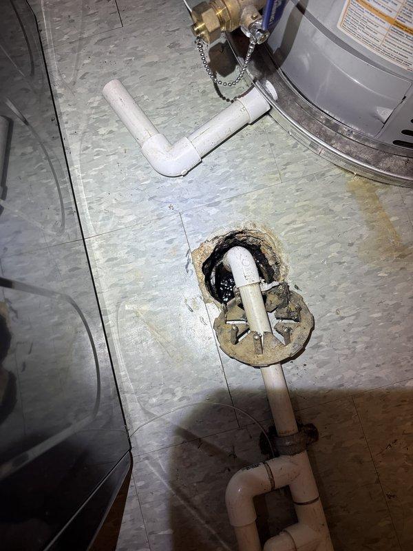 Responded to a laundry room floor drain backup with sewage overflow and inspected the drain line serving the floor drain. Exposed the floor drain connection, removing the corroded drain flange and broken concrete surround to access the PVC drain piping and metallic coupling, which contained heavy tar-like sludge buildup. Cleared the obstruction and cleaned out the debris, leaving the PVC assembly and fittings open for repair of the damaged drain connection.