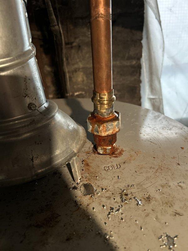 Responded to a reported leak at a union connection from a recent repipe and inspected the copper supply piping at the water heater. Found heavy corrosion and mineral buildup at the brass fitting and nipple on the cold water inlet, with rust staining on the tank top and dampness near the flue collar. Isolated the water supply and addressed the leaking connection to restore a watertight seal.