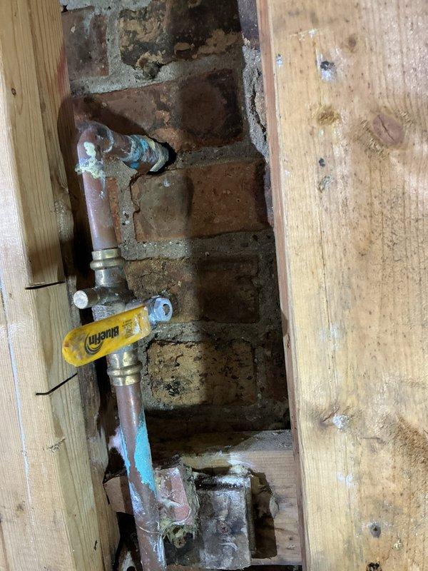 Assessed the kitchen supply piping to quote installation of a dedicated dishwasher shut-off valve. Inspected the existing copper supply line and in-line brass ball valve at the masonry wall penetration, noting corrosion, mineral buildup, and verdigris staining at the elbow and joints, along with a capped copper stub. Evaluated the cramped, cluttered work area and prepared the estimate for the independent isolation valve.