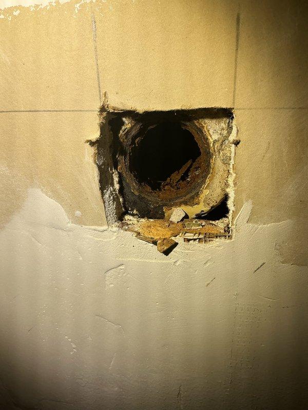 Accessed the in-wall plumbing at the repair location and documented the existing conditions, including multiple pipe penetrations through the drywall and a cut-out exposing a heavily corroded cast iron fitting with flaking rust and debris buildup. Marked layout reference lines on the wall for the planned repair work and surveyed the surrounding drywall and joint compound finish to assess the scope of the repair.