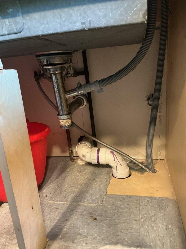 Responded to a clogged kitchen sink where prior homeowner attempts to clear the P-trap were unsuccessful. Inspected the drain assembly beneath the sink, including the tailpiece, slip-joint connections, PVC P-trap, and horizontal run to the wall, and cleared the blockage to restore proper drainage. Noted heavy buildup and corrosion on the brass coupling nut at the tailpiece, along with the dishwasher drain hose connection at the branch fitting, and verified the drain assembly was reassembled and flowing freely.