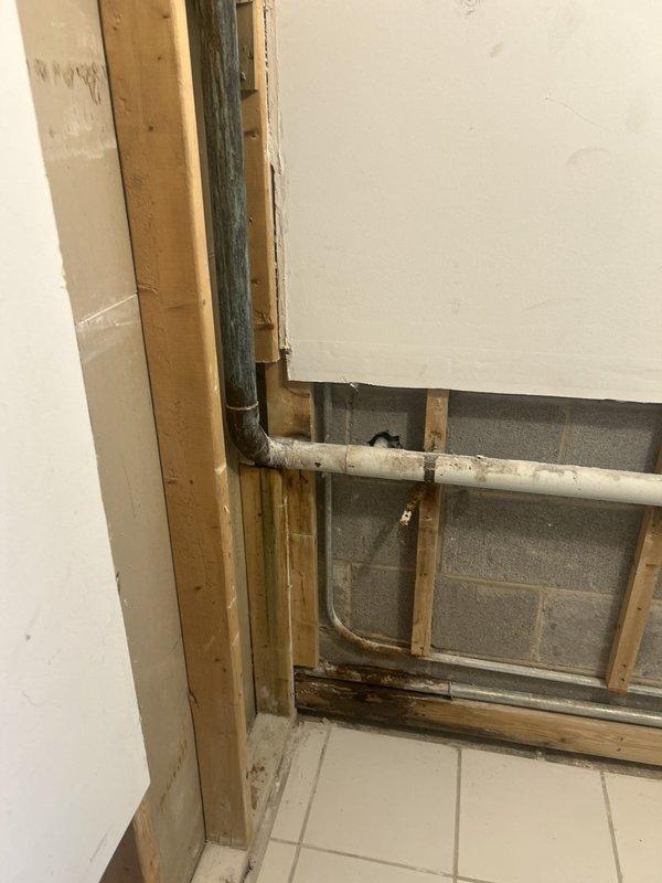 Opened the wall section by cutting away drywall to expose the framing and concrete block wall, gaining access to the existing dark supply piping transitioning into weathered white horizontal piping. Inspected the piping, strap supports, and water-stained bottom plate for leaks and moisture damage, and assessed the main shutoff valve and water meter assembly in the adjacent utility space as part of the repair.