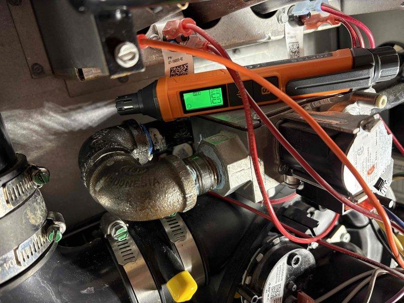 Responded to a persistent gas odor complaint and inspected the furnace gas train, including the black iron elbow and threaded connection to the gas valve, where corrosion and mineral residue were observed. Used a digital combustible gas detector to check the fittings, rubber couplings, and valve assembly for leakage and verify the source of the odor.