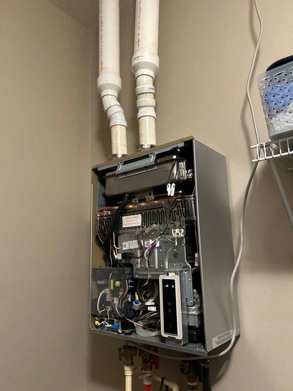 Performed assessment of Ruud tankless water heater (approximately 2 years old) that customer reports is approved for warranty replacement due to recurring issues. Visual inspection of the unit revealed intact internal components including circuit boards, wiring harnesses, heat exchangers, and proper gas line connections. Documented current installation configuration including PVC venting system, water line connections, and gas supply to facilitate proper reinstallation of warranty replacement unit when received from supplier.