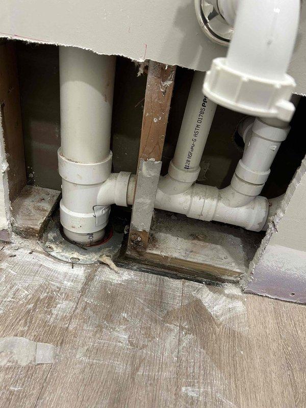Located and inspected cracked drain pipe behind bathroom sink wall. Accessed plumbing system through wall opening, revealing PVC drainage pipes with visible mineral buildup around connections. Repaired damaged section and verified proper drainage function with water supply on.