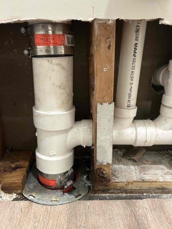Located and inspected cracked drain pipe behind bathroom sink wall. Accessed plumbing system through wall opening, revealing PVC drainage pipes with visible mineral buildup around connections. Repaired damaged section and verified proper drainage function with water supply on.