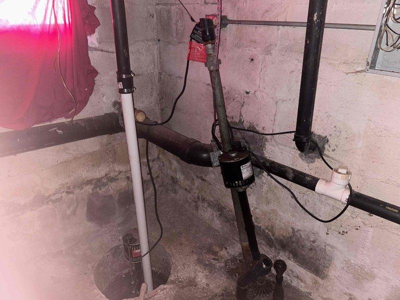 Conducted site assessment for sump pump relocation project in basement area. Documented existing plumbing configuration, including horizontal black pipe system connected to PVC components and current pump mounting location. Photographed the concrete block wall installation area and identified necessary connection points for the proposed relocation work.