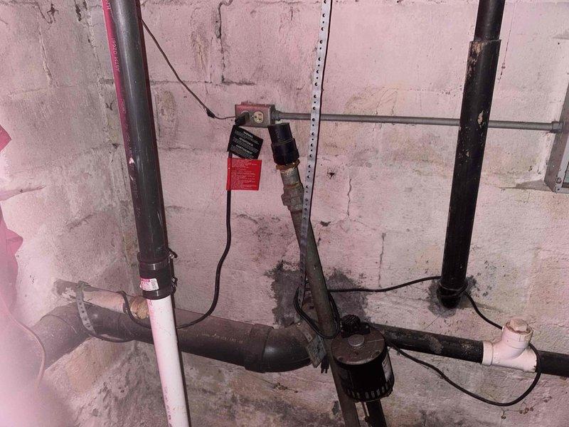 Conducted site assessment for sump pump relocation project in basement area. Documented existing plumbing configuration, including horizontal black pipe system connected to PVC components and current pump mounting location. Photographed the concrete block wall installation area and identified necessary connection points for the proposed relocation work.