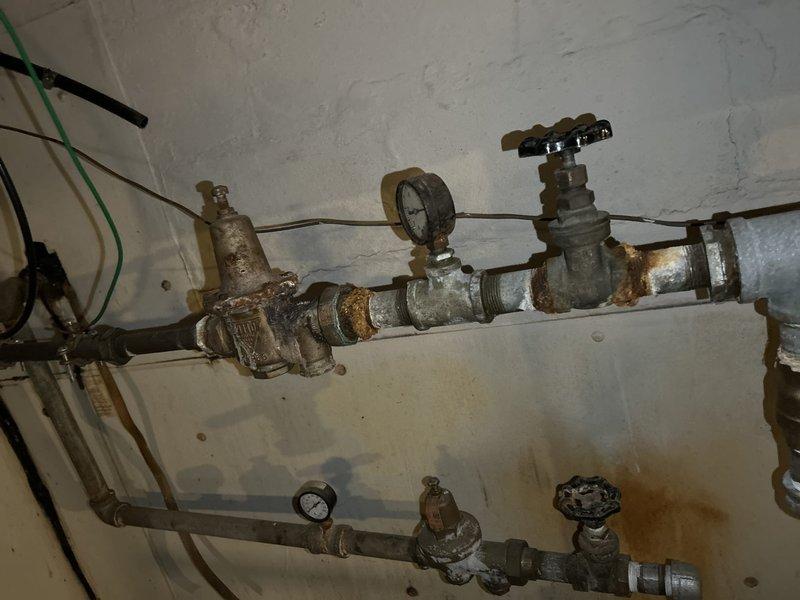 Conducted plumbing inspection of aging pipe system with significant corrosion and rust at joints and connection points. Documented extensive oxidation on metal components, particularly around pressure gauge assembly and valve fittings. Observed water staining on adjacent wall and insulated pipe section with mineral deposits, recommending system maintenance to prevent potential leaks and component failure.