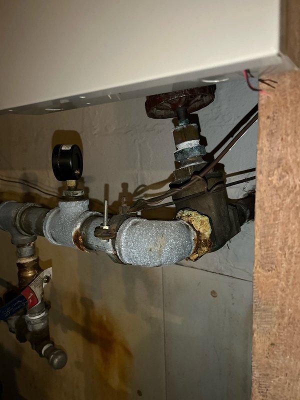 Conducted plumbing inspection of aging pipe system with significant corrosion and rust at joints and connection points. Documented extensive oxidation on metal components, particularly around pressure gauge assembly and valve fittings. Observed water staining on adjacent wall and insulated pipe section with mineral deposits, recommending system maintenance to prevent potential leaks and component failure.