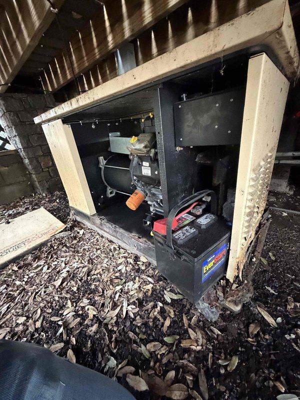 Performed routine maintenance on a Generac residential generator located under wooden patio behind house. Inspected control panel which showed normal operation with AUTO mode engaged and no warning indicators illuminated. Noted generator environment contained significant leaf debris accumulation around unit and battery, requiring cleaning to maintain proper ventilation and prevent potential fire hazards.