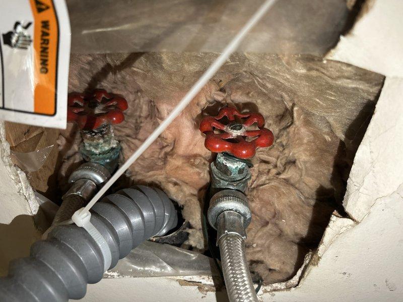 Conducted PSA plumbing inspection, examining water shutoff valves showing signs of oxidation within insulated wall cavity and shower control valve system. Documented condition of braided metal flexible connectors, pipe insulation, and valve functionality. Verified proper installation of bathroom fixtures including shower control valve and hose mounted on tiled wall surface.