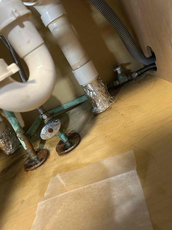 Conducted a plumbing inspection focusing on sink installation, noting oxidation and corrosion on copper/brass valves with improvised foil tape connections at joints. Observed water leakage at the base of the crystal handle faucet as evidenced by standing water on the sink surface.