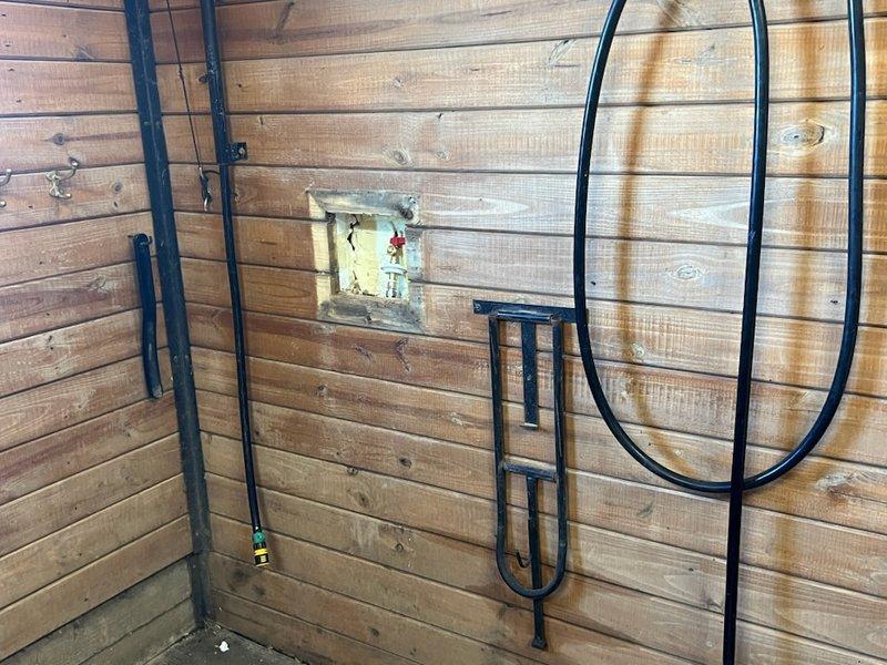 Inspected barn plumbing system where customer requested connection of existing lines. Identified a recessed water valve fixture with brass valve and red shut-off handle showing signs of deterioration and moisture damage in the wooden frame. Connected and secured the black metal pipes mounted on the wooden wall structure, ensuring proper integration with the existing junction box and brass fitting for functional outdoor water access.