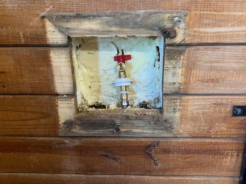 Inspected barn plumbing system where customer requested connection of existing lines. Identified a recessed water valve fixture with brass valve and red shut-off handle showing signs of deterioration and moisture damage in the wooden frame. Connected and secured the black metal pipes mounted on the wooden wall structure, ensuring proper integration with the existing junction box and brass fitting for functional outdoor water access.