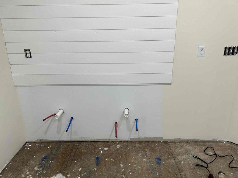 Completed plumbing estimate for bathroom rough-in with two sets of hot/cold water supply lines protruding from the wall, marked with appropriate color indicators. Observed electrical work in progress with multiple outlet boxes installed and noted preparation for future fixture installations including two circular wall cutouts requiring coordination with electrical components.