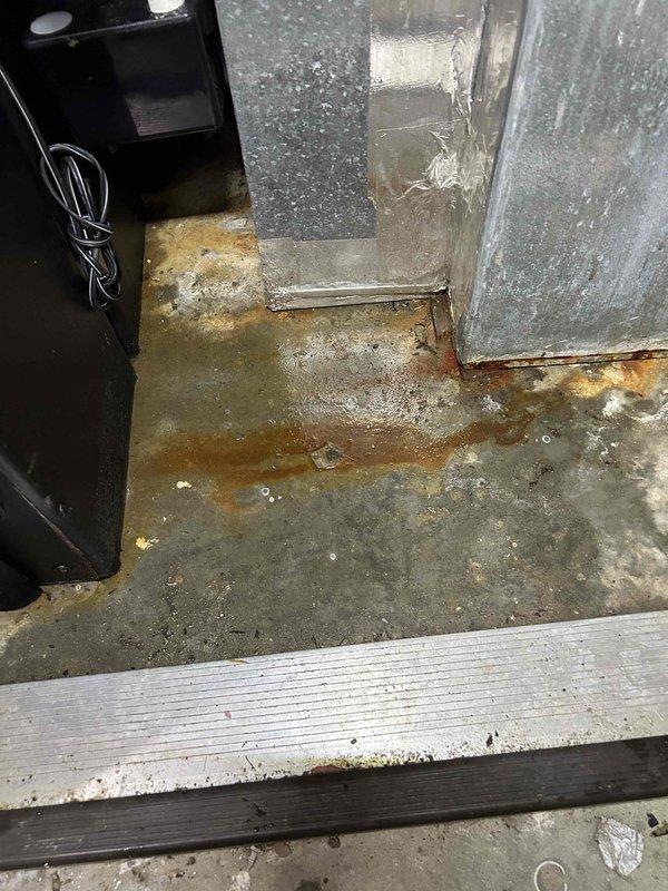 Responded to a no-cooling service call for an electric HVAC system under 10 years old. Discovered significant water damage and rust staining on the concrete floor beneath the air handler due to condensate drain line issues, with visible water leakage from the system. Identified that the filter was missing after previously being improperly secured with tape, potentially having fallen into the lower portion of the system, which was contributing to the operational failure and condensation problems.