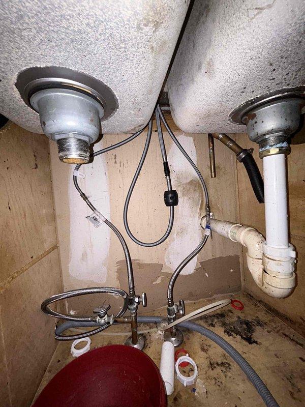 Conducted on-site assessment for kitchen sink installation. Observed existing double-basin sink with significant water damage and possible mold in the cabinet below. Noted deteriorated wooden cabinet floor with water collection basin present, suggesting ongoing leak issues. Examined current plumbing configuration including braided supply lines and PVC drainage components for replacement planning.