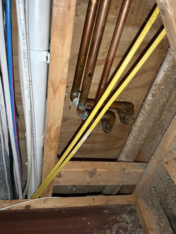Responded to plumbing service call for multiple leak issues in a 26-year-old residential property. Located and inspected leaking copper pipe running through floor/ceiling cavity between structural levels, documenting significant corrosion and rust deterioration on galvanized metal components with visible moisture staining and mineral deposits. Investigated reported kitchen sink leak, identifying actual source as separate issue beneath sink rather than sink fixture itself. Accessed confined structural space containing multiple utility systems including copper water lines, PVC drainage, and various conduits to assess extent of pipe deterioration and water damage. Photographed corroded sections showing compromised galvanized coating and active moisture infiltration points for repair documentation and quote preparation.
