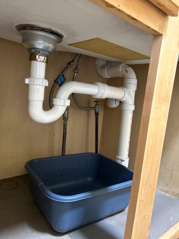 Completed installation of new sink drain assembly by drilling fresh hole in sink basin and installing chrome drain strainer with basket fitting. Connected white PVC P-trap configuration to new drain outlet and routed horizontal waste line to existing plumbing system. Installed new vent pipe running from drain assembly through cabinet and up through ceiling into basement area, ensuring proper drainage and venting per code requirements. All connections secured with appropriate fittings and tested for leaks. Verified proper drainage flow and vent function upon completion.