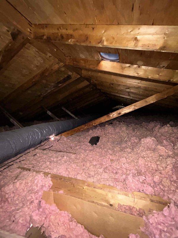 Responded to callback for air conditioning system not cooling, issue persisting from previous year's service attempt. Upon arrival, accessed attic space to inspect HVAC equipment and ductwork. Performed visual inspection of flexible duct system and surrounding installation conditions. Documented attic environment including exposed wooden framing, roof structure, and insulation coverage. Observed pink fiberglass insulation distributed across attic floor joists with gray flexible ductwork running through space. Noted general condition of attic components and captured photographic documentation of installation and environmental factors that may be impacting system performance for further diagnostic evaluation and repair planning.