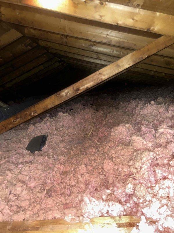 Responded to callback for air conditioning system not cooling, issue persisting from previous year's service attempt. Upon arrival, accessed attic space to inspect HVAC equipment and ductwork. Performed visual inspection of flexible duct system and surrounding installation conditions. Documented attic environment including exposed wooden framing, roof structure, and insulation coverage. Observed pink fiberglass insulation distributed across attic floor joists with gray flexible ductwork running through space. Noted general condition of attic components and captured photographic documentation of installation and environmental factors that may be impacting system performance for further diagnostic evaluation and repair planning.