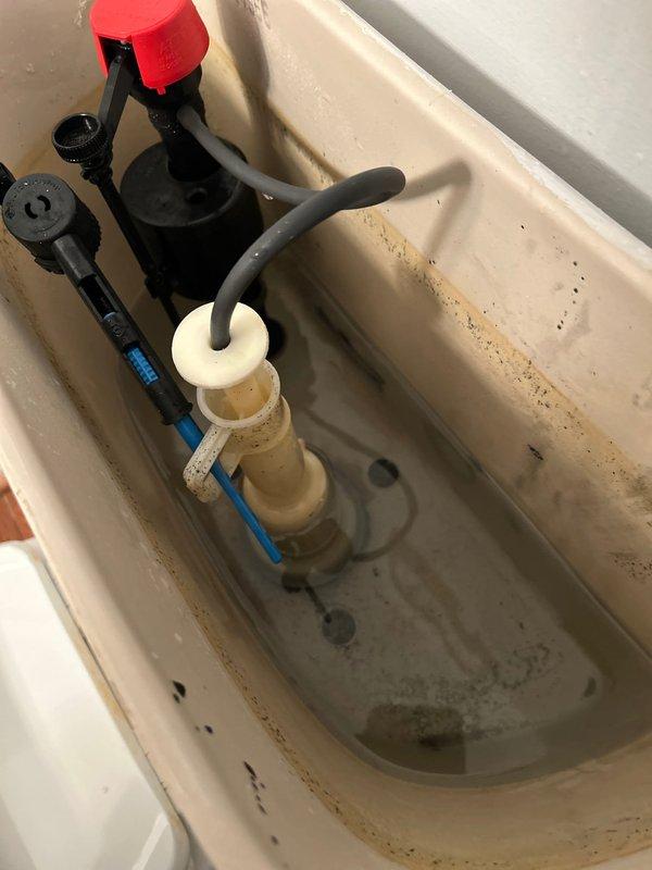 Performed routine plumbing inspection of toilet system, examining internal tank components including fill valve assembly, flush valve mechanism, flapper chain, and refill tube connections. Documented mineral buildup and water staining throughout tank interior, and verified proper installation and structural condition of toilet base and floor mounting.
