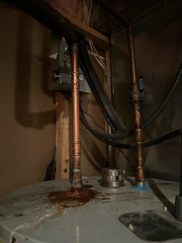 Responded to service call for leaking water heater fitting at top of tank. Upon arrival, confirmed active leak at copper pipe compression fitting connection where supply line enters water heater. Inspection revealed significant corrosion and mineral buildup at the joint, with rust staining and water damage visible on tank surface. The fitting showed deterioration consistent with previous leak history documented in account photos. Repaired the leaking compression fitting by disassembling the connection, cleaning corrosion from mating surfaces, and reinstalling with new compression ferrule and proper torque specifications. Verified all connections were secure and leak-free. Tested system under full operating pressure and confirmed no further leakage at repair site or surrounding connections. Cleaned work area and documented repair with photographs for service records.