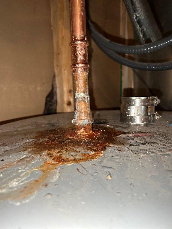 Responded to service call for leaking water heater fitting at top of tank. Upon arrival, confirmed active leak at copper pipe compression fitting connection where supply line enters water heater. Inspection revealed significant corrosion and mineral buildup at the joint, with rust staining and water damage visible on tank surface. The fitting showed deterioration consistent with previous leak history documented in account photos. Repaired the leaking compression fitting by disassembling the connection, cleaning corrosion from mating surfaces, and reinstalling with new compression ferrule and proper torque specifications. Verified all connections were secure and leak-free. Tested system under full operating pressure and confirmed no further leakage at repair site or surrounding connections. Cleaned work area and documented repair with photographs for service records.