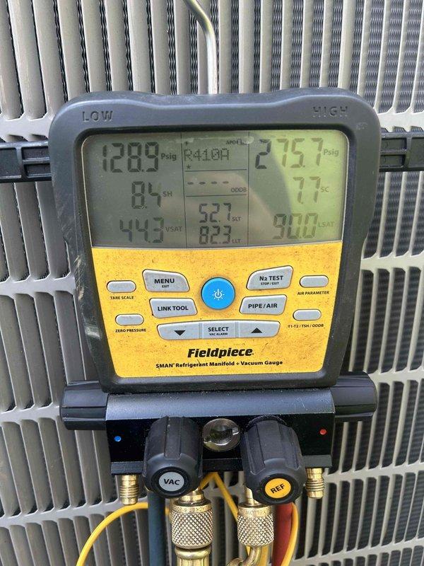 Performed electronic leak search on HVAC system using Fieldpiece SMAN refrigerant manifold and vacuum gauge. Connected diagnostic equipment to R-410A system and monitored refrigerant pressures, recording 128.9 psig on low side and 276.7 psig on high side at 84°F ambient temperature to identify leak location within the condenser unit.