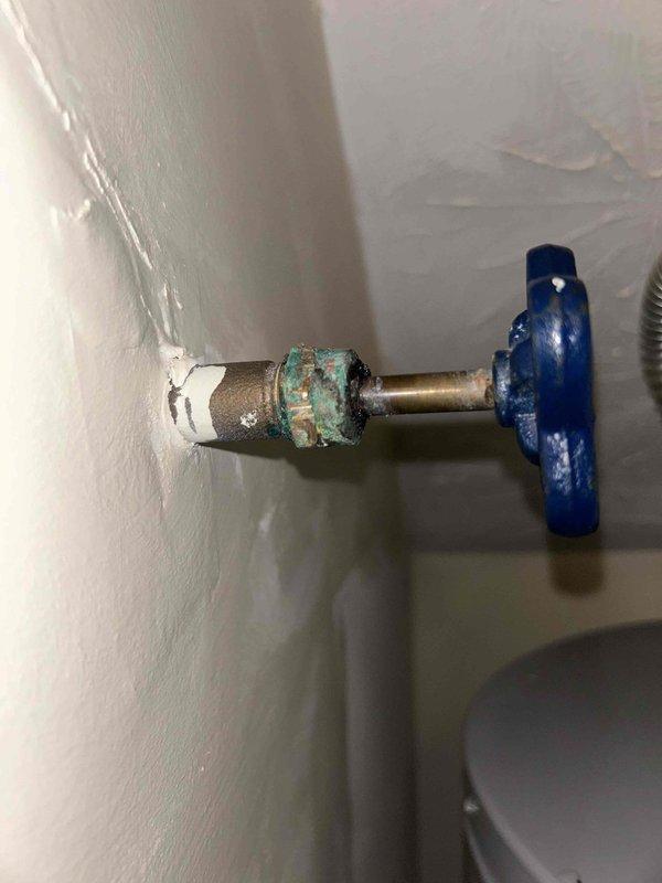 Responded to water leak complaint causing damage to flooring. Upon inspection, identified source as corroded shutoff valve with heavy verdigris buildup on brass section, indicating prolonged moisture exposure and oxidation. The valve assembly showed deterioration at connection points between white plastic wall mount, brass middle section, and blue plastic handle valve. Documented condition of affected plumbing fixture and surrounding water heater installation with AO Smith unit. Photographed corroded valve assembly and overall plumbing configuration including copper piping, PVC connections, and flexible relief hose for service records and future reference.