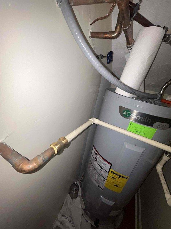 Responded to water leak complaint causing damage to flooring. Upon inspection, identified source as corroded shutoff valve with heavy verdigris buildup on brass section, indicating prolonged moisture exposure and oxidation. The valve assembly showed deterioration at connection points between white plastic wall mount, brass middle section, and blue plastic handle valve. Documented condition of affected plumbing fixture and surrounding water heater installation with AO Smith unit. Photographed corroded valve assembly and overall plumbing configuration including copper piping, PVC connections, and flexible relief hose for service records and future reference.