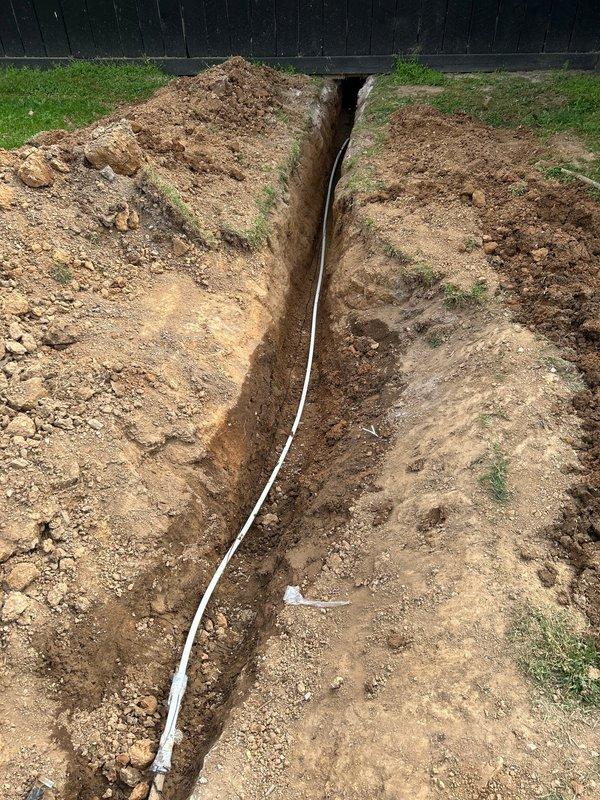 Excavated approximately 15 feet of water line from the meter to locate and access the existing supply line using an excavator. Dug interior trench inside barn structure through dirt floor to establish routing for new hydrant installation. Installed white PVC supply line running the length of the exterior trench and connected new yard hydrant at terminus point near barn structure.