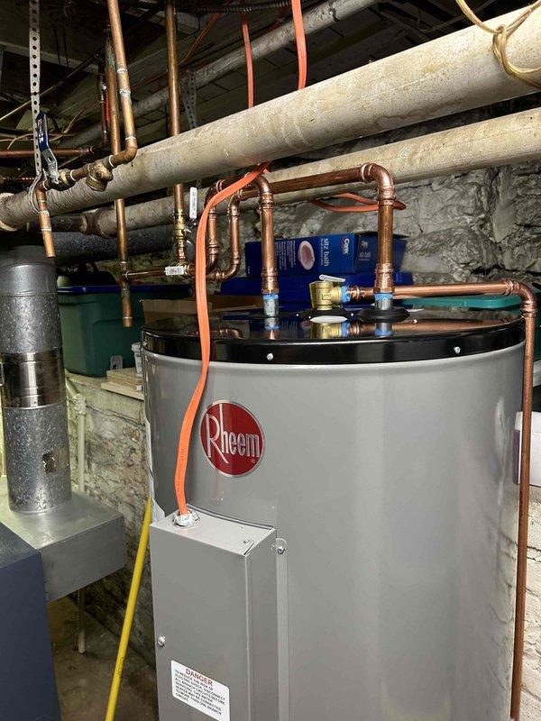 Removed existing 80-gallon electric water heater from basement utility area and installed new Rheem 80-gallon tanked water heater as replacement unit. Positioned new heater on elevated white metal platform stand to ensure proper clearance and accessibility for future maintenance. Connected unit to existing electrical supply, completing all wiring terminations at terminal block with proper color-coded conductors and secure connections within junction box enclosure.

Tied into existing hot and cold water supply lines using copper piping with soldered joints to ensure leak-free connections. Integrated new water heater with existing recirculation system, installing blue recirculation pump assembly on top of unit with appropriate copper pipe connections to maintain continuous hot water delivery throughout the building. Secured all horizontal supply lines to ceiling joists using proper pipe hangers and orange support straps to prevent sagging and ensure system longevity.

Completed all plumb