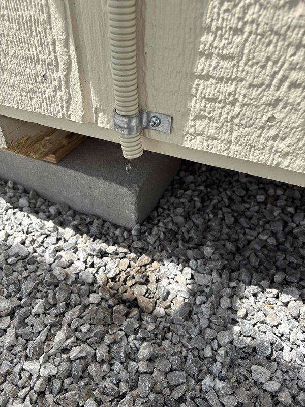 Completed installation of customer-supplied mini split system with outdoor unit mounted on ground-level bracket secured to building exterior wall. Installed electrical whip with conduit run along wall surface, coordinated with electricians for disconnect box mounting and final electrical connections. All refrigerant line sets and electrical components properly secured and protected, with unit positioned on gravel base for drainage and accessibility.