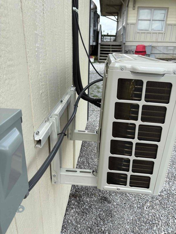 Completed installation of customer-supplied mini split system with outdoor unit mounted on ground-level bracket secured to building exterior wall. Installed electrical whip with conduit run along wall surface, coordinated with electricians for disconnect box mounting and final electrical connections. All refrigerant line sets and electrical components properly secured and protected, with unit positioned on gravel base for drainage and accessibility.