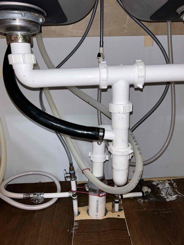 Responded to clogged drain line affecting dishwasher operation. Upon inspection of the under-sink plumbing assembly, identified blockage in the drainage system connected to the dishwasher discharge line. Cleared obstruction from the drain line using appropriate methods, ensuring proper water flow through the P-trap assembly and main drainage piping. Verified all connections, including the black rubber dishwasher discharge hose secured with metal hose clamps to the white PVC drainage system, were properly seated and functioning. Tested system operation to confirm complete drainage and no leaks present at any connection points.